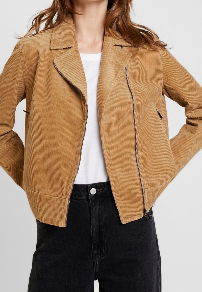 Beżowa kurtka z kordurowego materiału o skróconym kroju, z wyciętym kołnierzem, zamkiem błyskawicznym oraz bocznymi kieszeniami, w połączeniu z białą bluzką i czarnymi spodniami.