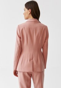 Růžový blazer s přiléhavým střihem, strukturovanými rameny a zádovými záševky. Hladká látka s jemným leskem, doplněná výstřihovým límcem.