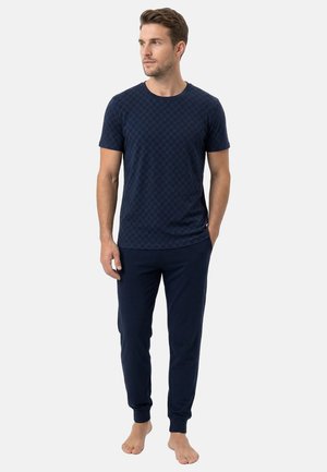 Man staat op blote voeten, draagt een navyblauw T-shirt met korte mouwen en patroon en bijpassende navyblauwe joggingbroek.