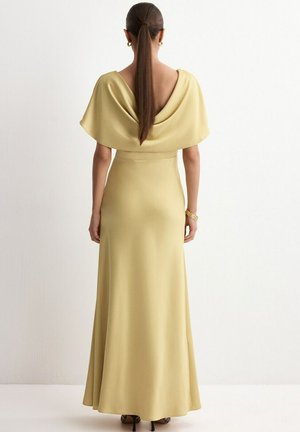 Kobieta z długim kucykiem, ubrana w sięgającą podłogi bladożółtą suknię z odkrytymi plecami i złotą bransoletką, stojąca na tle białej ściany.