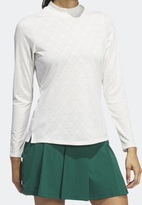 Bílé tričko s dlouhým rukávem a jemným geometrickým vzorem s vysokým límcem, doplněné zelenou plisovanou sukní. Obě části jsou vyrobeny z hladké látky.