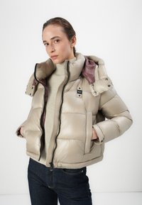 Beige pufferjack met een glanzende afwerking, grote capuchon, voorrits en zijzakken. Gelaagd over een lichtgekleurde geribde trui.