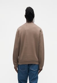 Καφέ φούτερ με στρογγυλή λαιμόκοψη και λεπτομέρειες ριμπ μανσέτας, σε συνδυασμό με τζιν παντελόνι ανοιχτού γαλαζί χρώματος. Ορατές ραφές και άνετη, χαλαρή εφαρμογή.