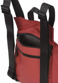 Mochila de tela roja con correas negras ajustables y un bolsillo frontal con cremallera. Presenta una textura suave y un diseño minimalista.