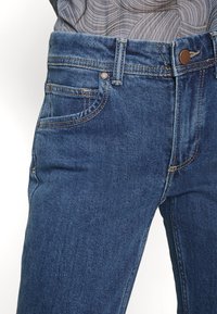 Denim jeans in dark blue with a classic five-pocket design, featuring brown hardware, a button closure, and visible stitching details.