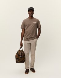 Brun t-skjorte med "LES DEUX" tekst, lysebeige bukser, mørke sko og en rutete duffelbag. Kappen har et rutenett mønster.