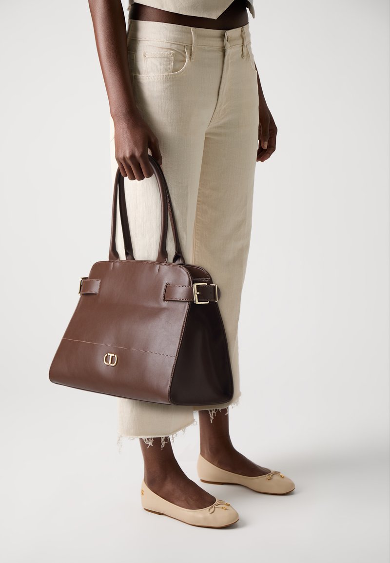 Brun læderhåndtaske med en struktureret design, dobbelt håndtag, sidelukninger og en guldspænde. Sammen med cremefarvede jeans og beige flade sko.
