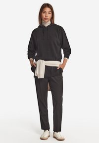 Schwarzer Kapuzenpullover mit einem weißen Rollkragenpullover darunter, schwarze Hose, cremefarbener Pullover um die Taille gebunden und weiße Sneakers.