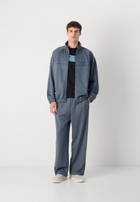 Herreidrettsantrekk i slate-grått, bestående av en jakke med glidelås og elastiske mansjetter, matchende løstsittende bukser og en svart T-skjorte med lyseblå detaljer.