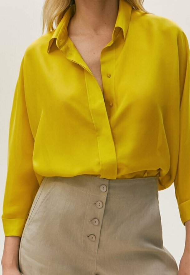 Femme portant une chemise boutonnée jaune vif rentrée dans un pantalon beige taille haute avec quatre boutons visibles.