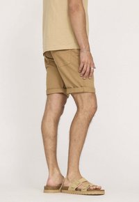 Shorts en coton beige avec des revers, des poches latérales et une coupe décontractée. Portés avec des sandales à enfiler beiges.