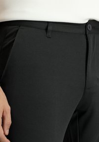 Pantaloni neri realizzati in materiale testurizzato, con frontale piatto, passanti per cintura e chiusura con un solo bottone. Dettaglio ravvicinato del tessuto.