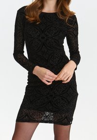 Frau in einem schwarzen Langarm-Minikleid mit filigranen Blumenmustern, die mit gefalteten Händen vor ihr steht.