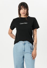 T-shirt noir en coton à manches courtes, arborant le logo blanc "Calvin Klein" sur la poitrine. Porté avec un pantalon en denim léger bleu clair taille haute.