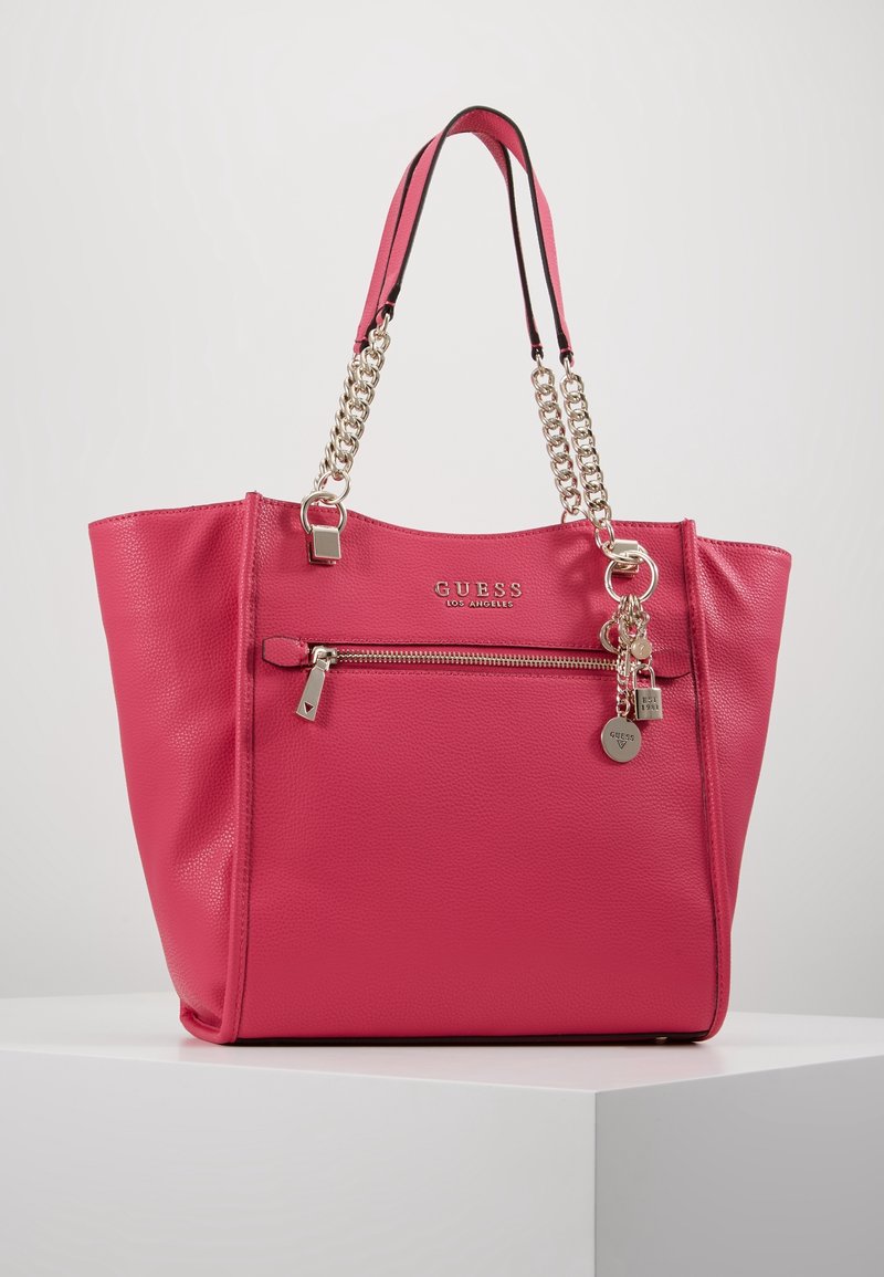 Sac à main Guess en cuir rose vif avec anses chaîne dorée, poche avant zippée, logo en métal et détails charmants sur une surface blanche.