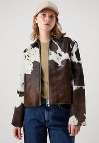 Lederjacke aus Kuhhaut mit einer Mischung aus braunen und weißen Mustern, ausgestattet mit einem Reißverschluss, einem spitzen Kragen und langen Ärmeln. Kombiniert mit einem einfachen Oberteil.