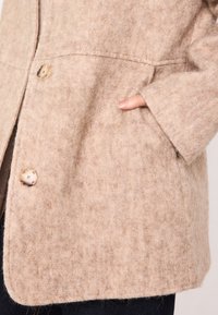 Cappotto in misto lana beige con una texture morbida, chiusura a bottoni a pressione e tasche laterali. Presenta una vestibilità rilassata e un orlo curvo.