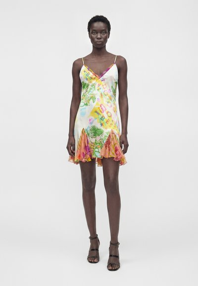 Γυναίκα που φοράει αμάνικο, πολύχρωμο μίνι φόρεμα με λουλούδια, βολάν στο τελείωμα και λεπτές τιράντες, συνδυασμένο με μαύρες ανοιχτές γόβες.