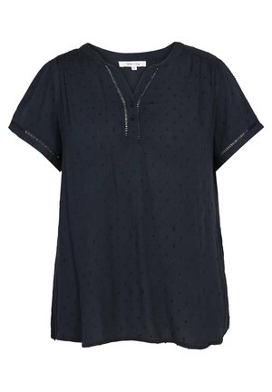 Kvinde iført en marineblå kortærmet bluse, lyseblå cropped jeans og hvide sneakers, stående med den ene hånd på hoften og den anden arm afslappet.