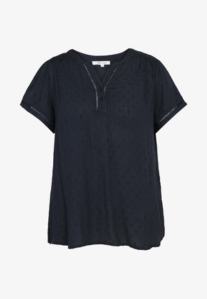Femme portant un chemisier bleu marine à manches courtes, un jean court bleu clair et des baskets blanches, debout avec une main sur la hanche et un bras détendu.