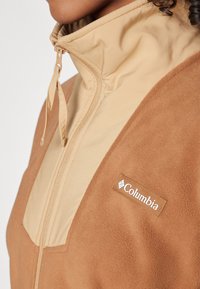 Brun fleecejacka med ljusbeiga detaljer, hög krage och framzipper. Har en Columbia-logotyp på sidan. Mjuk textur.