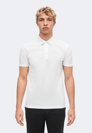 Ung mand med blondt krøllet hår iført en ensfarvet hvid kortærmet poloshirt og sorte bukser står mod en lys baggrund.