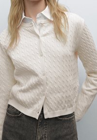 Weißer Strickcardigan mit V-Ausschnitt und Knopfleiste, verziert mit strukturierten Mustern. Getragen über einem weißen Hemd mit Kragen, kombiniert mit dunkler Jeans.