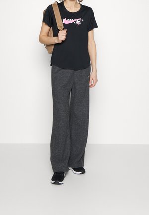Sort kortærmet t-shirt med et pink Nike-logo, parret med grå vide sweatpants og sorte sportsko. En lysebrun rygsæk hænger over den ene skulder.