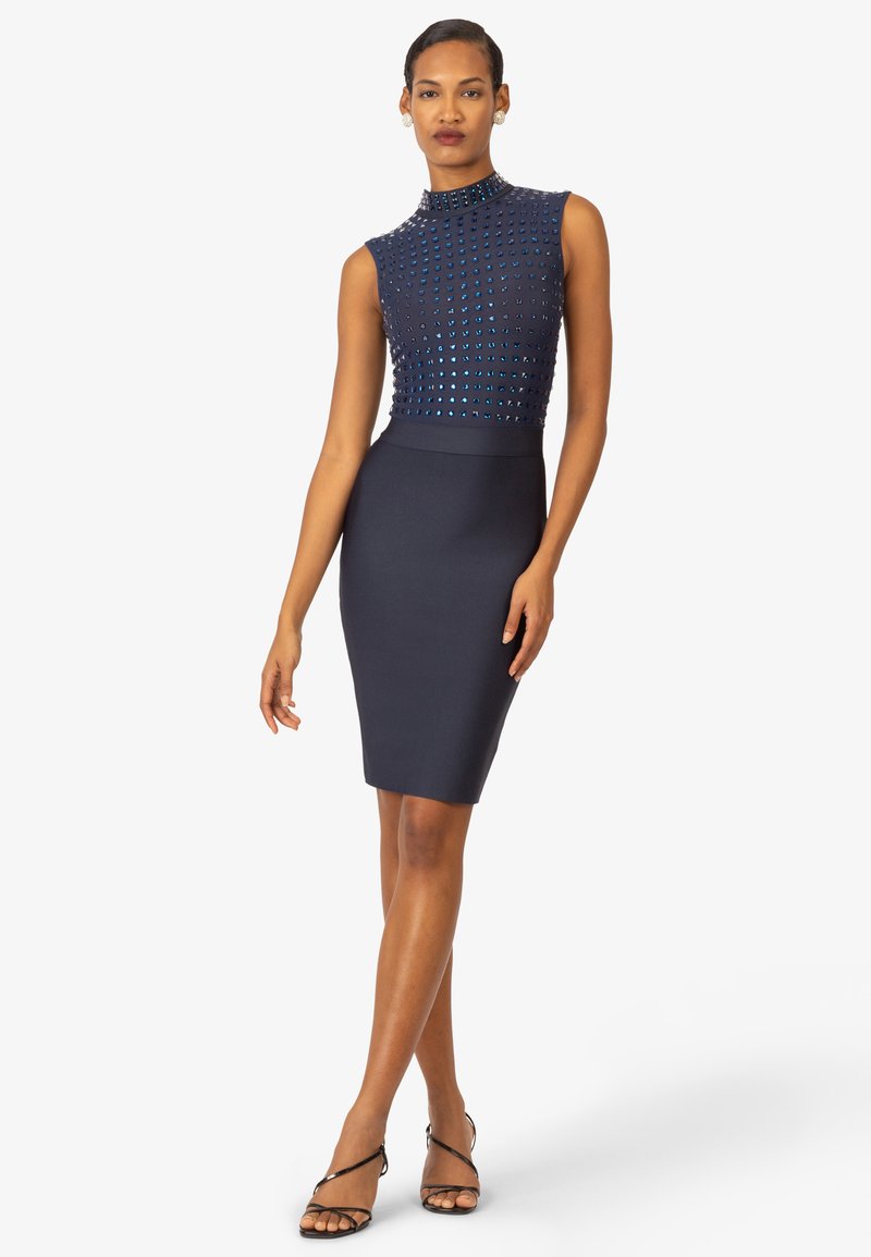 Marineblaues Kleid mit tailliertem Schnitt, das einen mit Nieten besetzten Oberteil und einen hohen Kragen aufweist. Der Bleistiftrock reicht bis zum Knie.