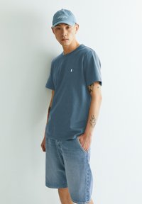 Blauwe katoenen t-shirt met een klein wit logo, gecombineerd met lichtblauwe denim shorts. Het model draagt een bijpassende blauwe pet.