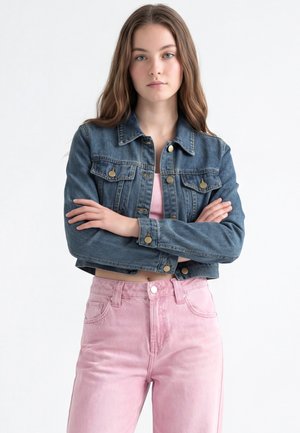 Junge Frau mit langen Haaren, die eine kurze Jeansjacke und hoch taillierte rosa Hosen trägt, steht mit verschränkten Armen vor einem weißen Hintergrund.