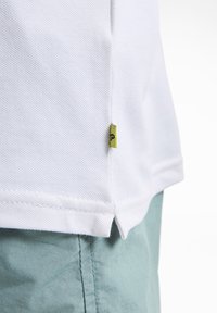 Polo shirt blanc en tissu texturé, avec une petite étiquette verte sur l'ourlet. Le shirt est associé à un short vert clair.