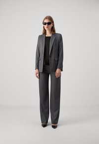 Suit grigio su misura con giacca strutturata, pantaloni a gamba larga e top nero. Il modello indossa occhiali da sole neri e tacchi a punta neri.