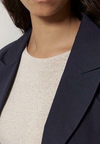 Blazer blu navy con revers affilati sopra un top in maglia di un beige chiaro e strutturato. Collo aderente e un mix di tessuti lisci e pregiati.