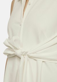 Witte mouwloze top met een knoopsluiting en een knoopdetail bij de taille, gemaakt van een gladde, lichte stof.
