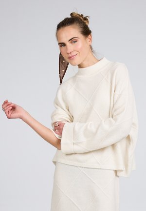 Frau mit braunem gepunktetem Schal im Haar trägt einen cremefarbenen Strickpullover und passenden Rock, posiert mit einem angewinkelten Arm und einem leichten Lächeln.