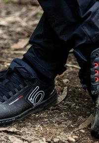 Scarpe sportive nere con una superficie testurizzata, caratterizzate da accenti bianchi e rossi, lacci e una suola robusta, posizionate su una superficie naturale.