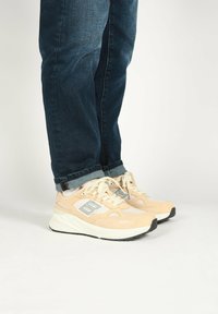Light peach athletic shoes with mesh and suede materials, featuring a white sole, reflective accents, and gray branding details. Paired with blue jeans.