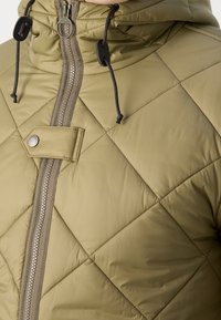 Gesteppte olivgrüne Jacke mit hohem Kragen, Frontreißverschluss und Zugkordel-Verstellungen. Auffälliger Metall-Druckknopf und strukturierte Oberfläche.