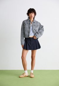 Jovem em pé sobre um chão verde, vestindo uma jaqueta cinza cropped, uma blusa listrada, uma saia azul marinho, meias brancas e ténis bege, com um fundo de parede branca.
