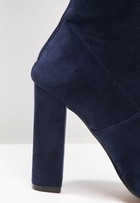 Bottine en suède bleue marine avec un talon bloc haut, présentant une texture lisse et un subtil détail de couture le long de la couture.