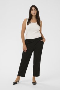 Haut blanc sans manches et pantalon noir sur mesure. Les jambes sont droites, le tissu est lisse. Chaussures noires à bout pointu avec des accents de nœud.