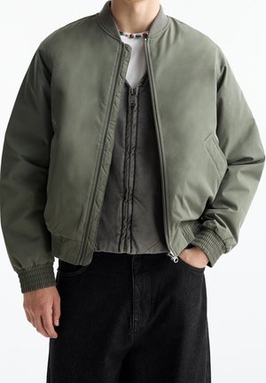 Mann trägt hellolivgrüne Bomberjacke über grauer Zip-Weste mit weißem Hemd, schwarze Hose, eine Hand in der Tasche vor weißem Hintergrund.