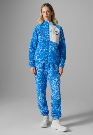 Vrouw die een blauw-wit fleecejack met patroon en broek draagt met witte sneakers, staand tegen een effen grijze achtergrond.