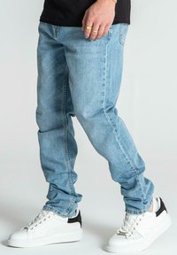 Jeans de denim azul claro con un corte relajado, que cuentan con bolsillos y una cremallera, combinados con zapatillas blancas que tienen acentos en negro.