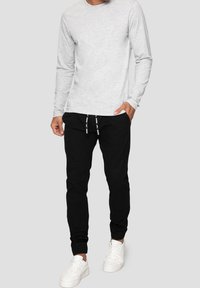 Man i ljusgrå långärmad skjorta, svarta joggerbyxor med dragsko och vita sneakers, som står mot en enkel bakgrund.