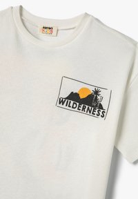Valkoinen ribattuja t-paita lyhyillä hihoilla, jossa musta ja keltainen "Wilderness" -graafinen painatus, joka esittää vuoria ja auringon.