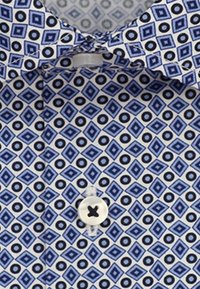 Modrá a bílá košile s vzorem diamantů a kruhů, zobrazující knoflík a část límce svázanou uzlem.