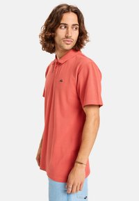 Herren Poloshirt mit kurzen Ärmeln, Korallenfarbe, aus Baumwollstoff, mit Knopfleiste am Kragen, kleinem Logo auf der Brust, lässiger Schnitt, glatte Textur.