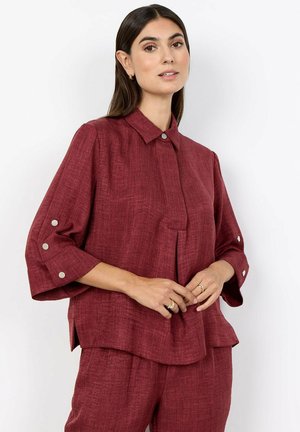 Vrouw met lang donker haar, gekleed in een loszittend, gestructureerd donkerrood overhemd met knopen en bijpassende broek, staand tegen een effen achtergrond.